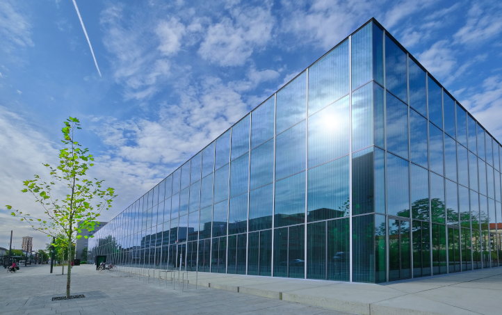 Quaderförmiges Museumsgebäude mit Glasfassaden, in denen sich Wolken und Sonne spiegeln, links daneben ein junger Laubbaum